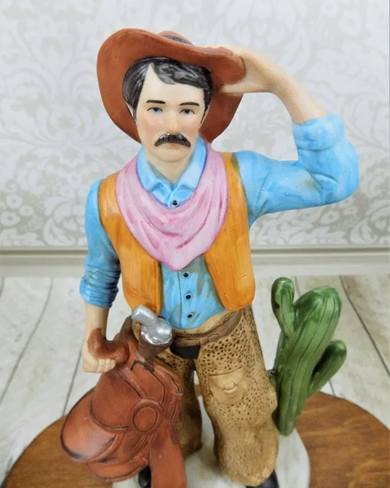 A vintage figurine of a dark-haired cowboy with a mustache wearing a brown hat, a blue shirt,an orange vest, and pink handkerchief, and holding a leather horse saddle.