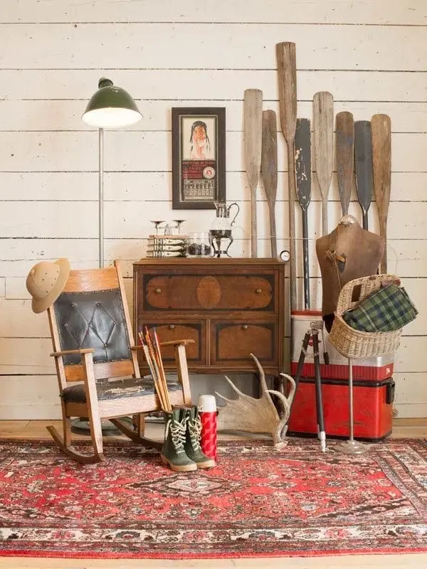 A vintage Wild West decor arrangement featuring a vintage rug, a wood and leather rocking chair, wooden dresser, vintage boat paddles, and other vintage decor items against a rustic cream-colored shiplap wall.