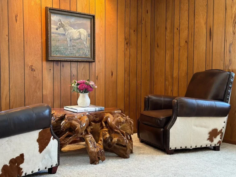 Two dark leather armchairs partially upholstered in cowhide stand on a white carpet, with a hand carved wooden coffee table, all-over wall paneling, and a vintage painting of a horse on the wall.