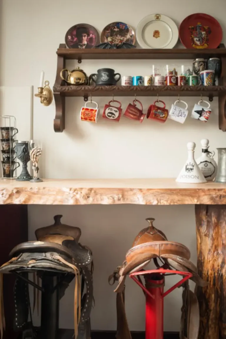 A vintage wood cupboard with vintage mugs, teacups, and plates hangs on a white wall above a rough wooden table top with vintage bar stools made from horse saddles.