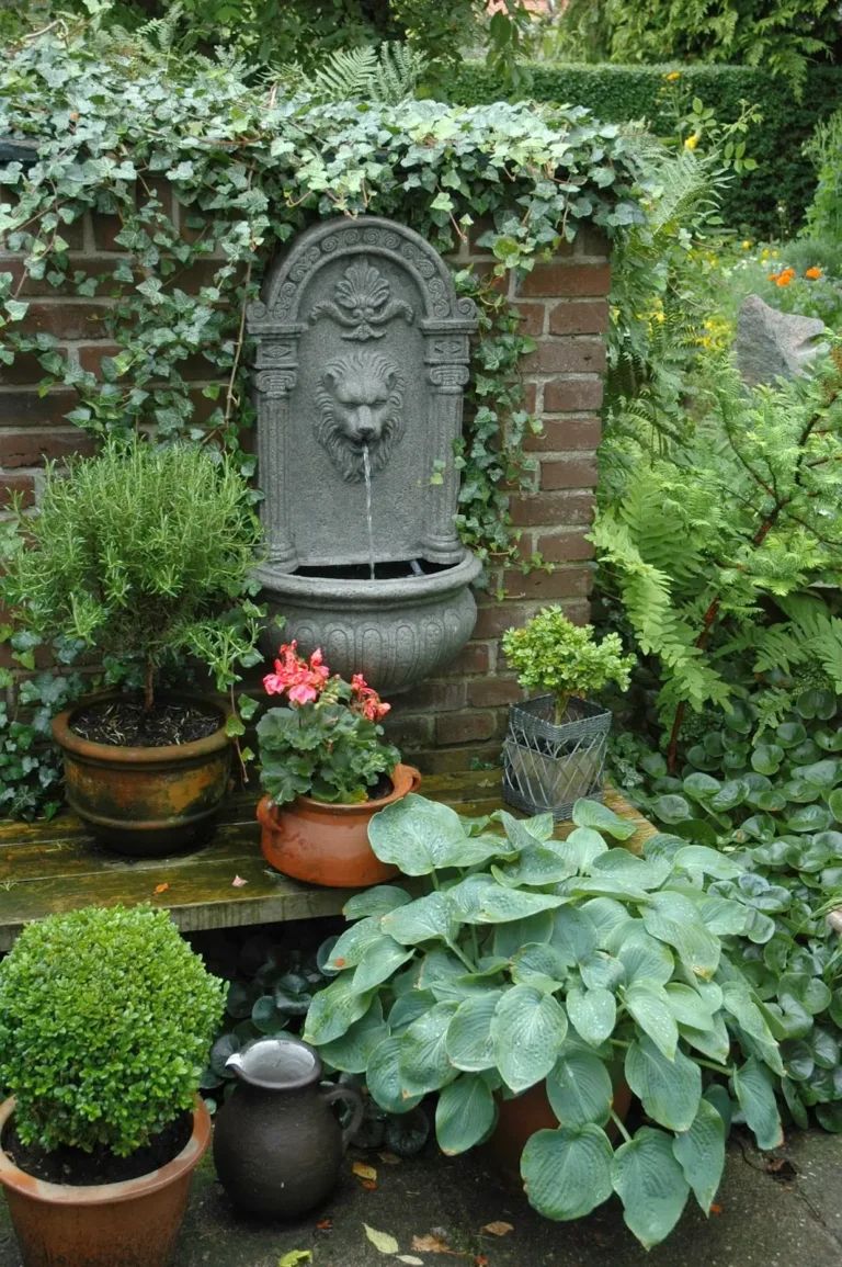 Ferns and other green plants in terracotta pots surround an antique-style gray garden fountain with the head of a lion.