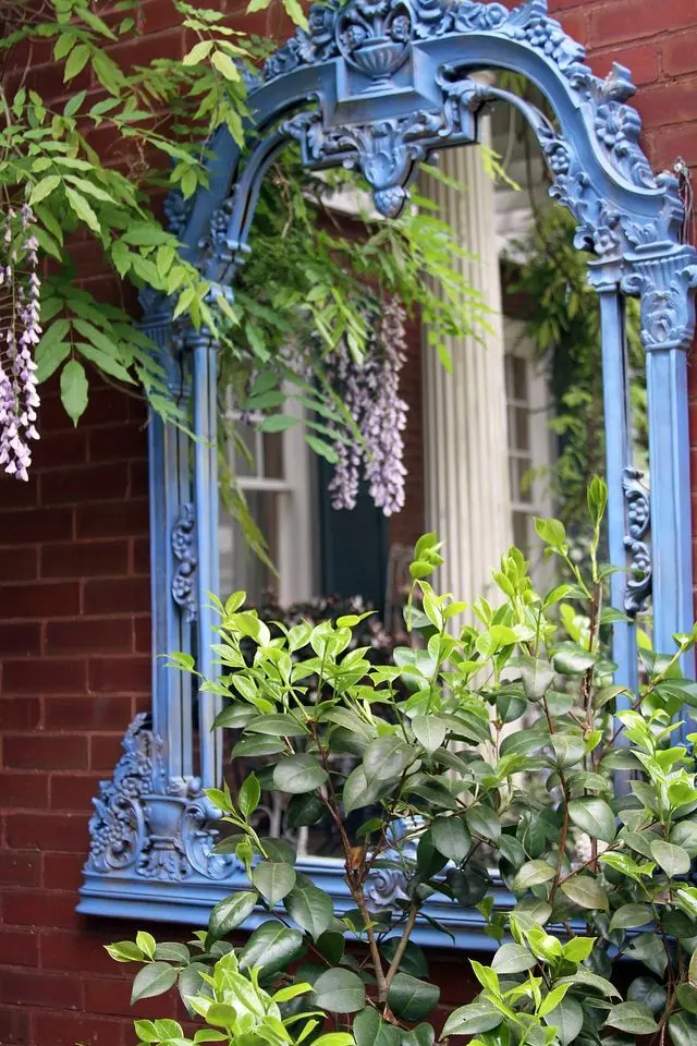 An intricately carved blue antique mirror hangs on a red brick all, with green plants in front of it.