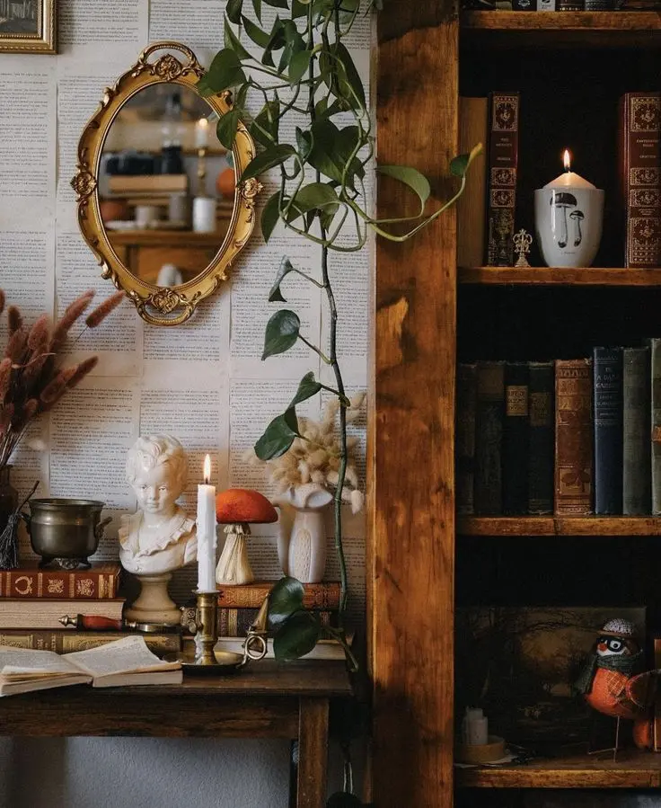 An oval mirror in an ornate gold frame hangs on a wall wallpapered with book pages. There’s a brown desk with antique books, a candelabra with a white candlestick, a vintage bust, figurine of a toadstool, and some other vintage items, and an old wooden bookcase next to it.