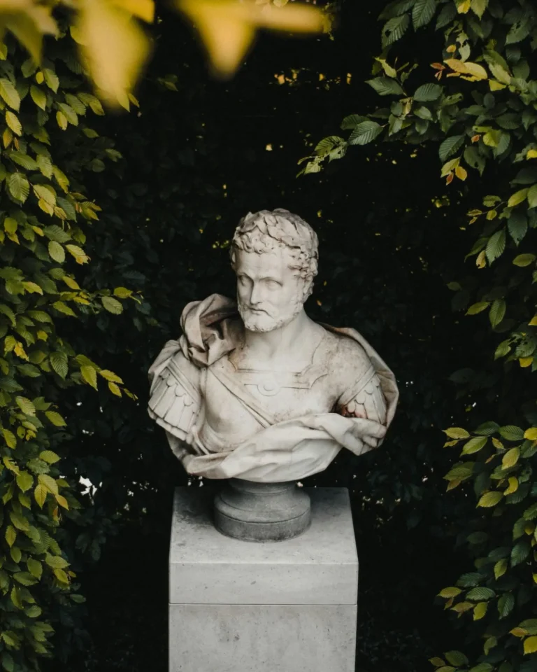 An antique Greek sculpture in white marble surrounded by shrubs of greenery.