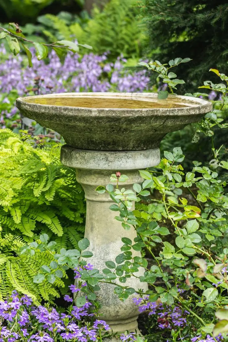 An antique bird bath surrounded by ferns, purple flowers, and other garden plants.