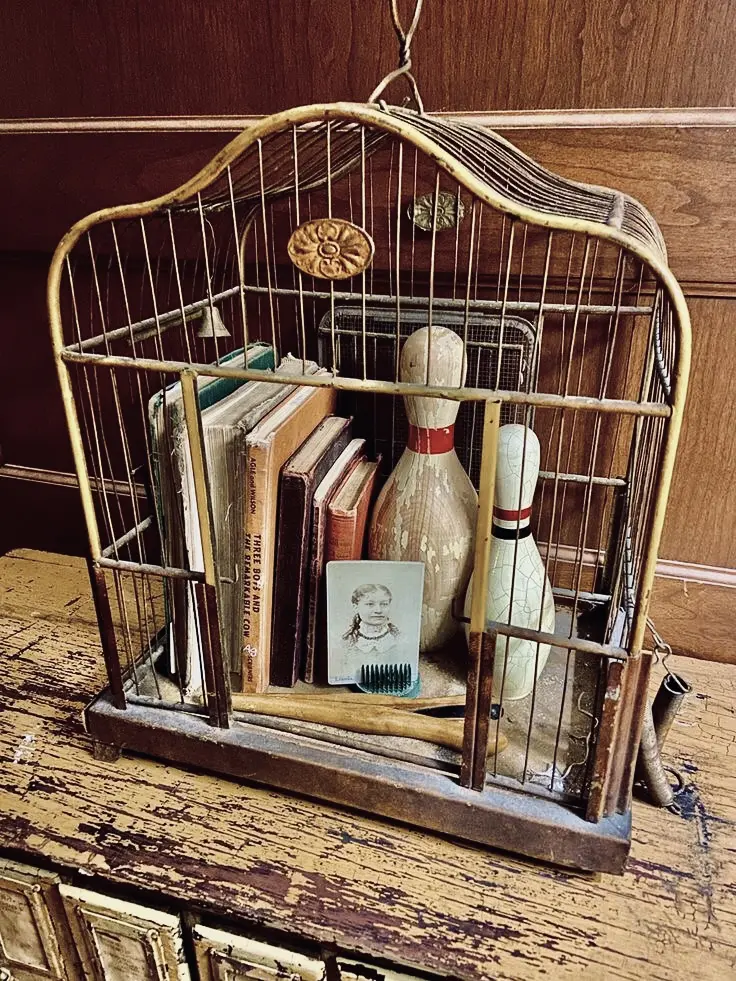 A couple of old books, two vintage bowling pins, and a 19th century daguerreotype inside a decorative worn out cage sitting on a rustic wooden piece of furniture.