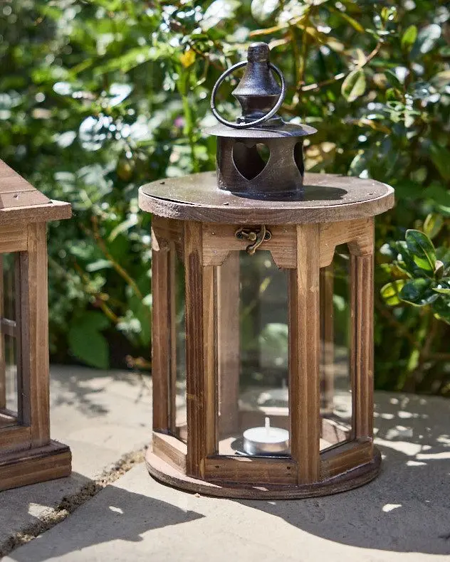 A hexagonal wooden lantern with a white tea light inside sits on a concrete slab, with green shrubs visible in the background.