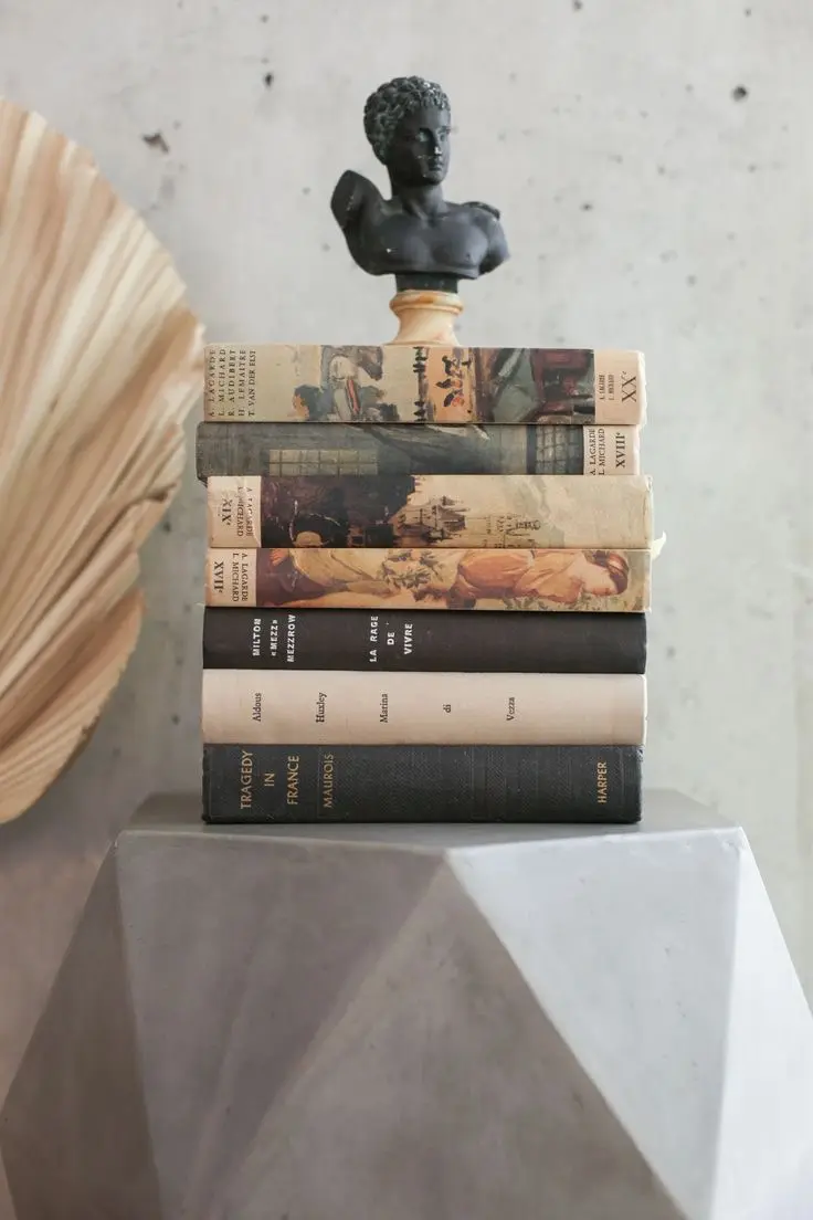 Seven beige and black light academia-style books stacked on top of each other, with a small dark Greek sculpture on top.