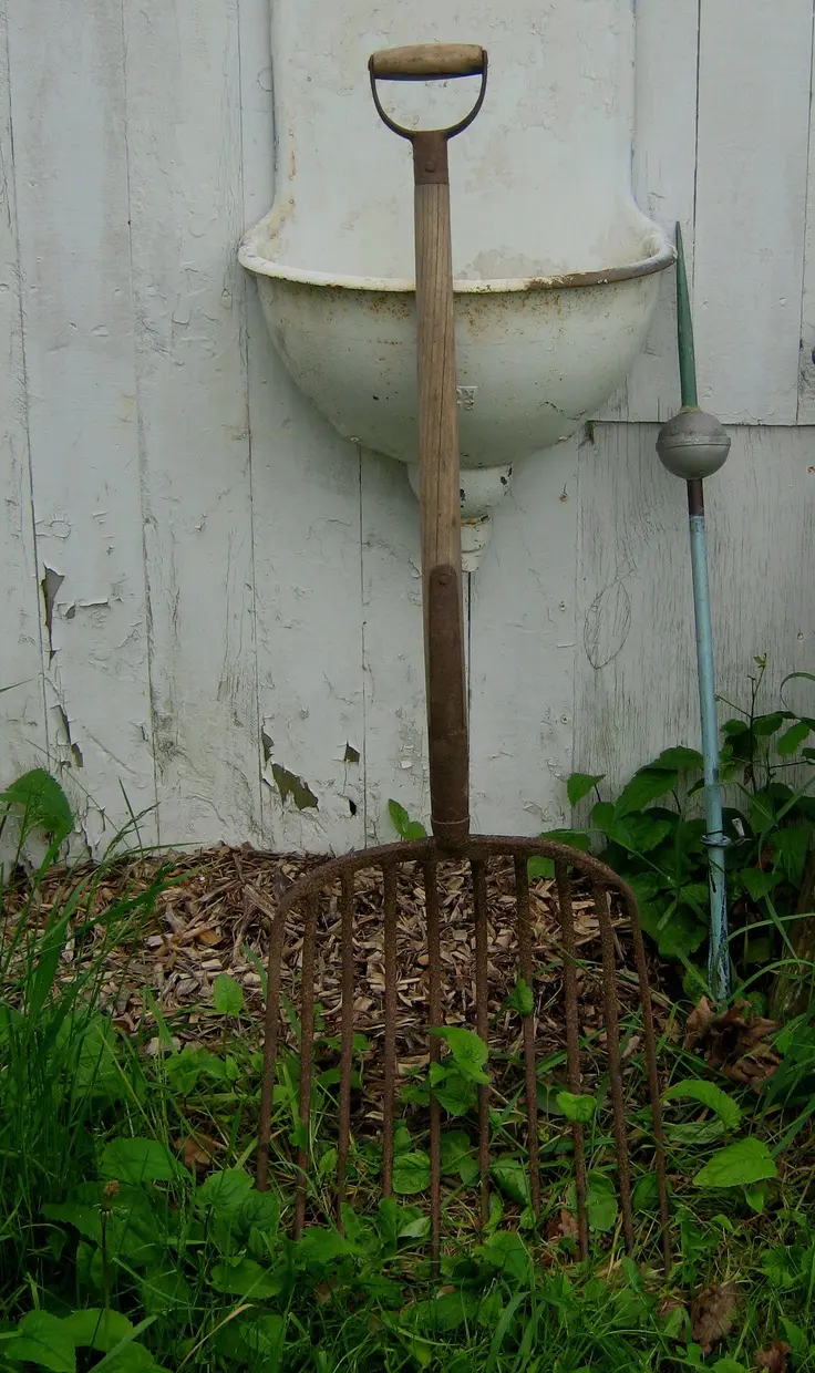 An old, white, half-oval garden sink is mounted on a distressed white paneled wall; a wide vintage garden pitchfork is leaning against the sink.