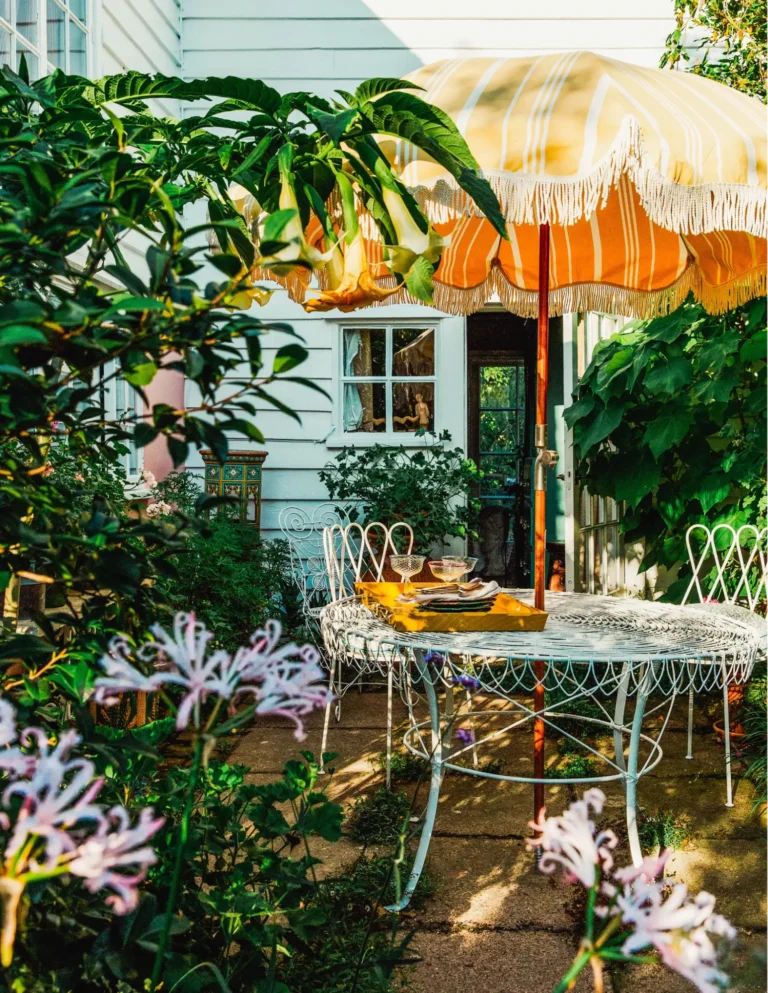 Vintage garden decor featuring a white metal table and chairs set, a vintage striped patio umbrella, and lots of green shrubs, all in front of a turquoise-panelled house.