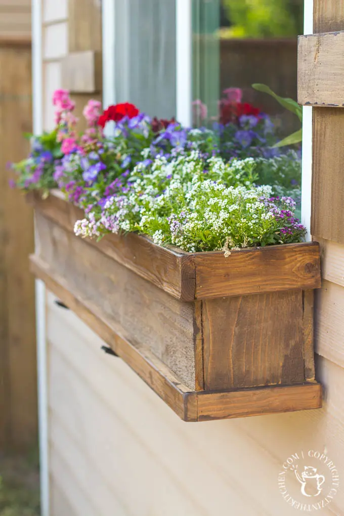 Colorful garden flowers planted in a rustic wooden window box hanging below a window on a cream-paneled home.