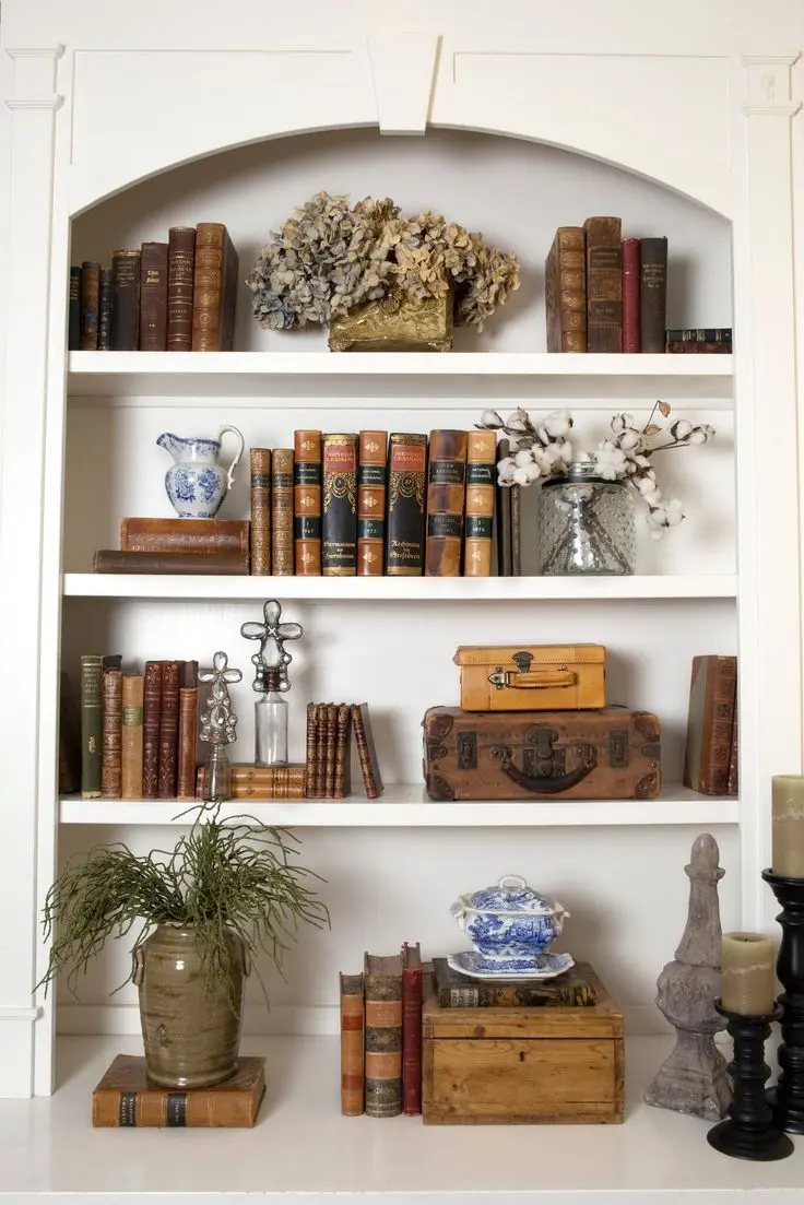 A classic white bookshelf with three bookshelves and an arched top, displaying antique books, blue and white china, candelabras, dried flowers, and a plant in a clay vase.