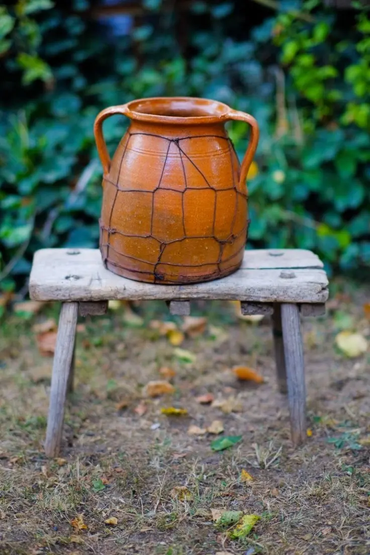 An orange-colored vintage ceramic jug sits on a small rustic wooden stool outdoors, with green shrubs seen in the background.