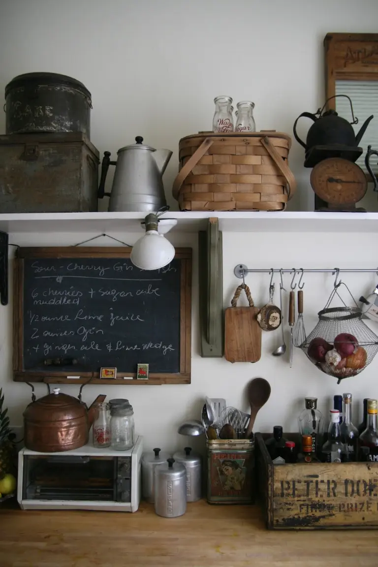 A wooden kitchen countertop with various vintage accessories such as a wooden crate, vintage utensils, baskets, a copper kettle, and a rustic chalkboard encased in a wooden frame.