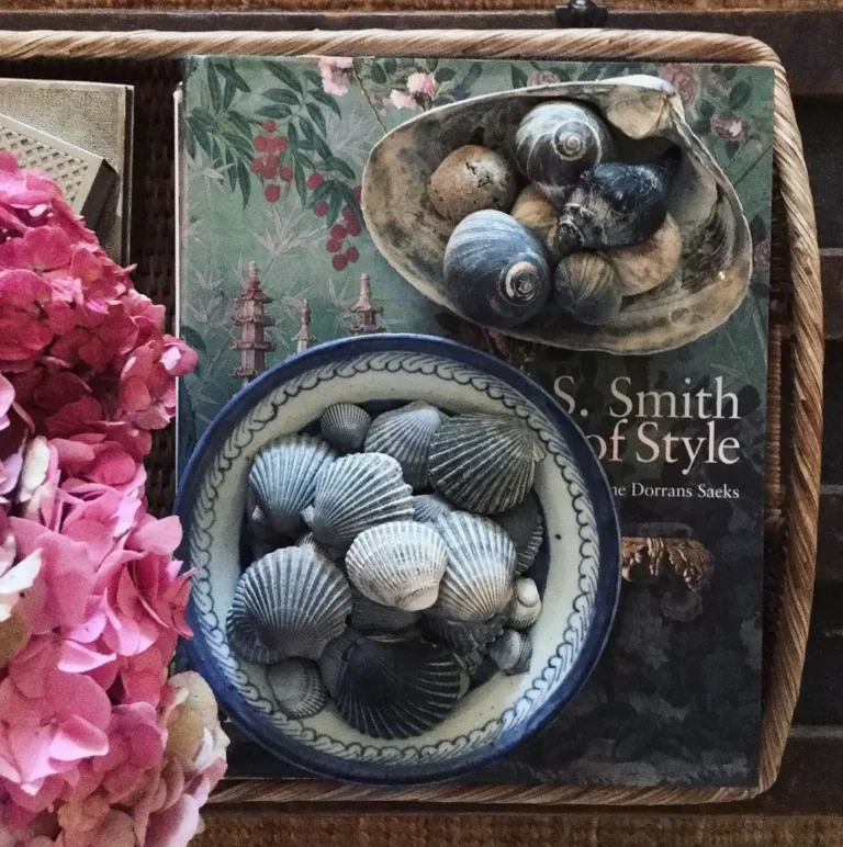 A vintage blue and white bowl filled with blue seashells and a large seashell filled with seashells sit on top of a large vintage-style coffee table book arranged on a tray, with pink flowers next to it.