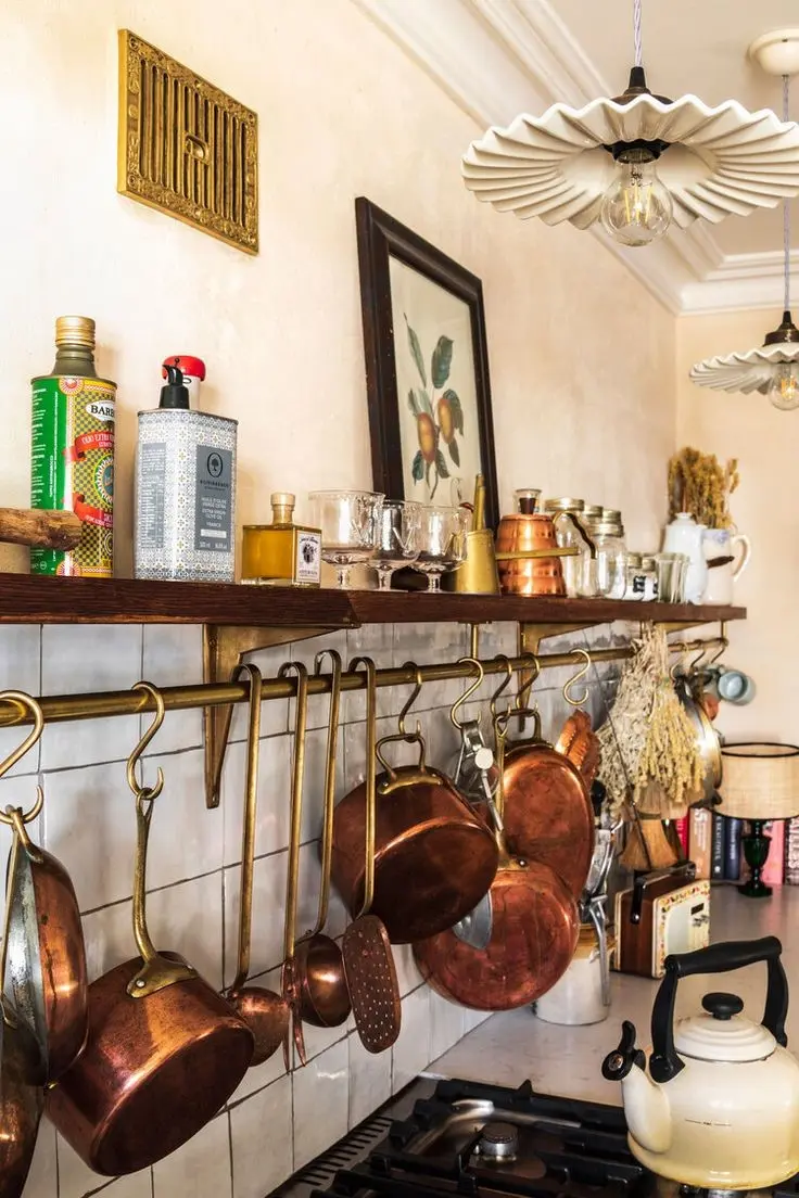 Vintage pans, pots, and other cookware hang on a brass bar on a white-tiled wall, surrounded by other vintage kitchenware and a vintage botanical poster in a black frame.