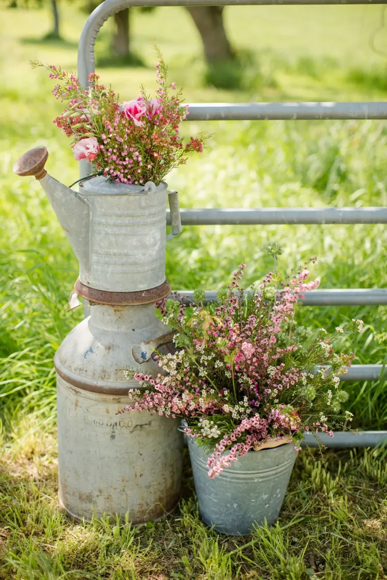 A rustic watering can sits on top of an old milk can, with a metal pail filled with a bouquet of wildflowers next to it.