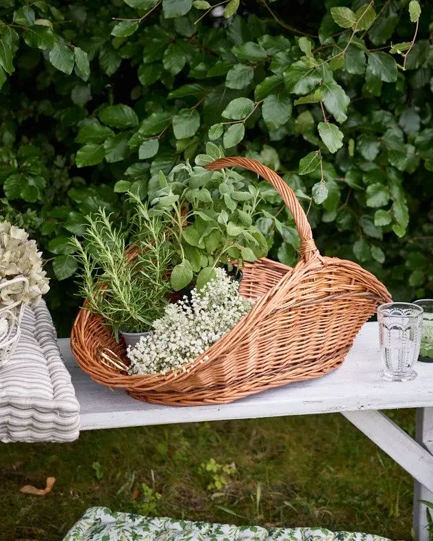 Two small pots of rosemary and mint and a small bouquet of white wildflowers sit inside a light brown wicker basket situated on a white rustic bench, with a green shrub behind it.