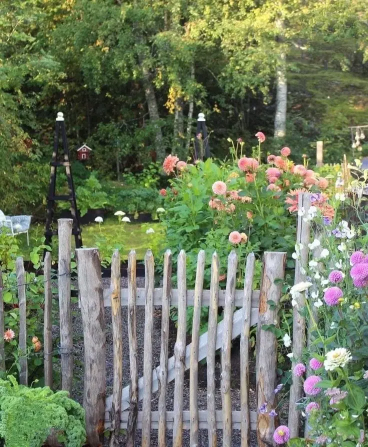 A rustic wooden gate surrounded by shrubs and colorful flowers, with more flowers, trees, and a lawn seen behind it.