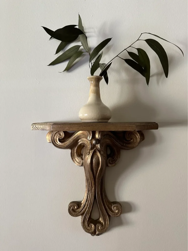 An antique bronze floating shelf hanging on a cream-colored wall and displaying a vintage beige vase with two plant cuttings.