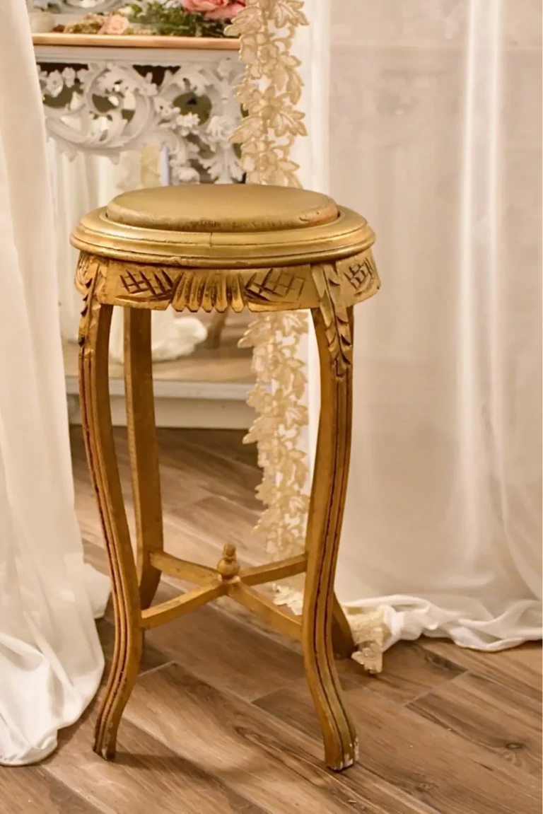 An antique gold-colored French stool on a wooden floor, with white decorative curtains in the back.