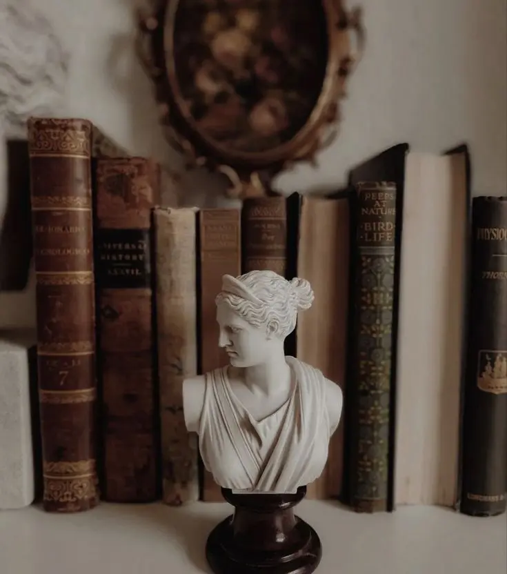 A small bust of the Greek goddess Diana on a small dark brown pedestal stands on a white surface with antique books and a botanical painting in an ornate oval frame in the background.