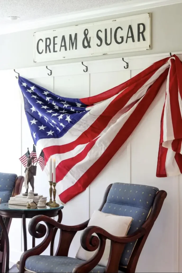 A draped American flag hanging from black wall hooks on a white paneled wall, a sign above it that reads Cream & Sugar, and a vintage rocking chair in the front.