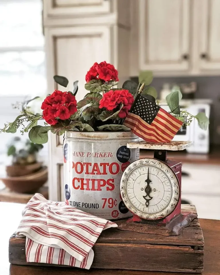 A vintage-inspired vignette made up of a rustic wooden tray decorated with a ticking striped linen, a vintage kitchen scale, and an old potato chips can with red geraniums and a vintage American flag.