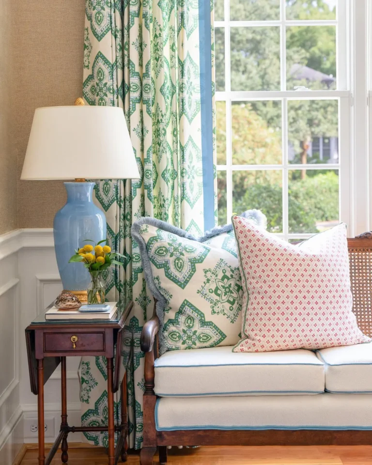 An example of grandmillennial decor featuring patterned pillows on a cane-back sofa with white, blue-trimmed cushions, a blue and cream table lamp on a cherry brown end table, and green, blue, and cream patterned drapes.