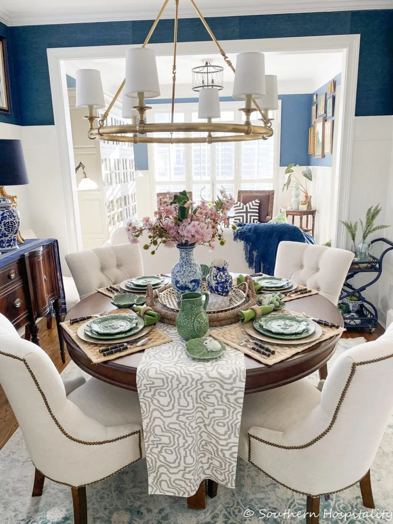 A grandmillennial dining room featuring a round mahogany table set for lunch, four cream-colored tufted chairs with brown wood legs, a bouquet of fresh pink flowers in a blue and white vase, and a blue-painted bamboo bar cart seen in the background.