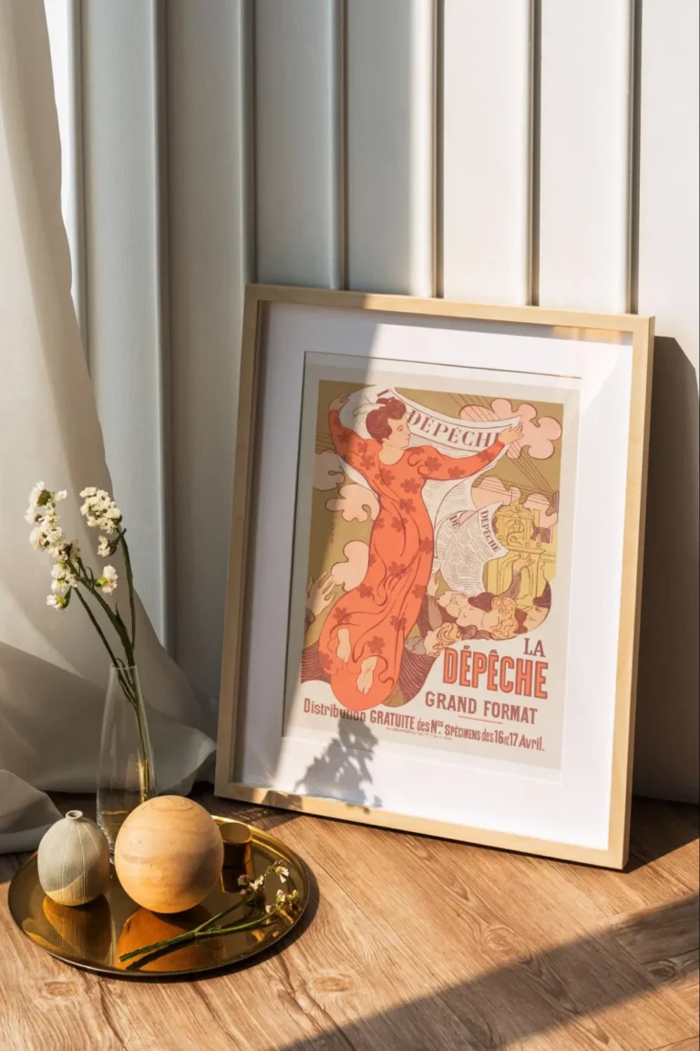 A framed Belle Epoque poster advertising the 19th century French newspaper La Depeche; next to it is a gold round tray with a small clay vase, a round sphere, and a glass vase with white flowers.