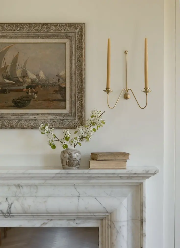 A nautical-themed oil painting in a decorative frame and an antique brass wall sconce with yellow candles hang on a white wall; below them is a marble fireplace mantel with flowers in a small vase and a stack of two books.