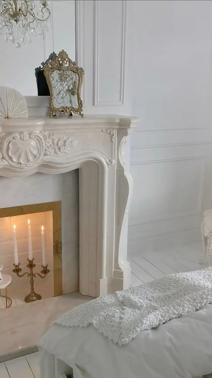 An ornately decorated white marble mantel with a brass candelabra with lit white candles inside. Other items in the photo include a crystal chandelier, a brass decorative frame, white floor panels, decorative wall moldings, and a white blanket and bedding.