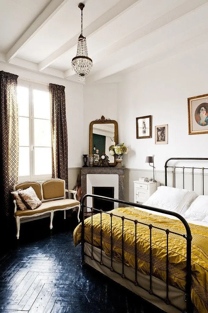 A vintage bedroom with a black herringbone wood floor, a black metal bed with a gold quilted bedspread, an antique sofa, a white bed stand, an ornate mirror, a chandelier, a fireplace, and artwork on the walls.