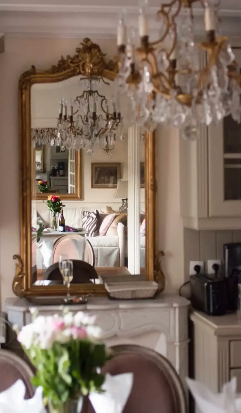 A rectangular ornate brass mirror on top of a marble mantel with some furniture and a chandelier seen in the reflection.