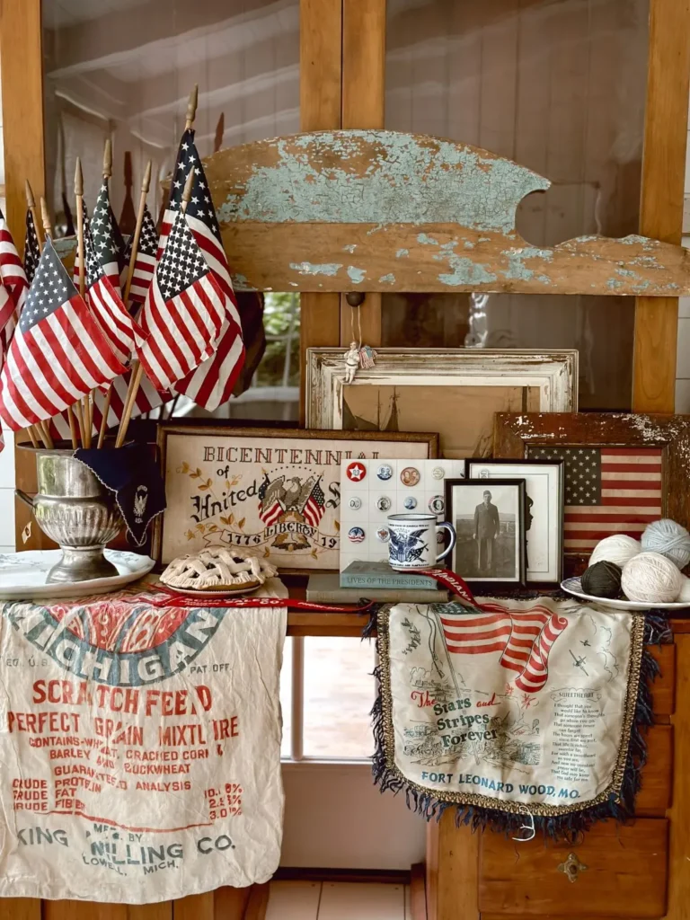 A vintage patriotic vignette featuring worn-out linens and textiles, American flags, a black and white photo of a veteran, vintage U.S. illustrations, rustic wooden frames, and an apple pie on a plate.