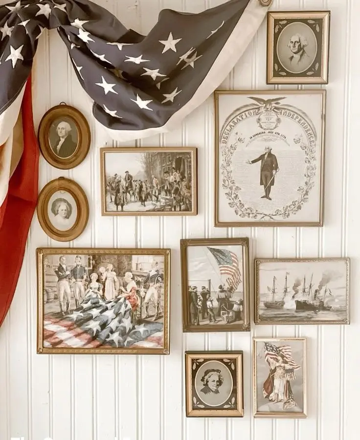 A gallery wall made up of vintage patriotic illustrations in retro frames hanging on a white beadboard wall with an old American flag draped on top.