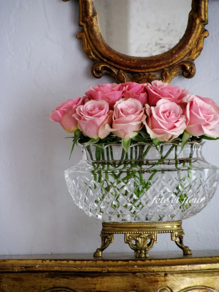 A bouquet of fresh pink roses in a beveled glass vase on ornate brass legs, sitting on top of an antique piece of furniture, with an ornate brass mirror in the background.