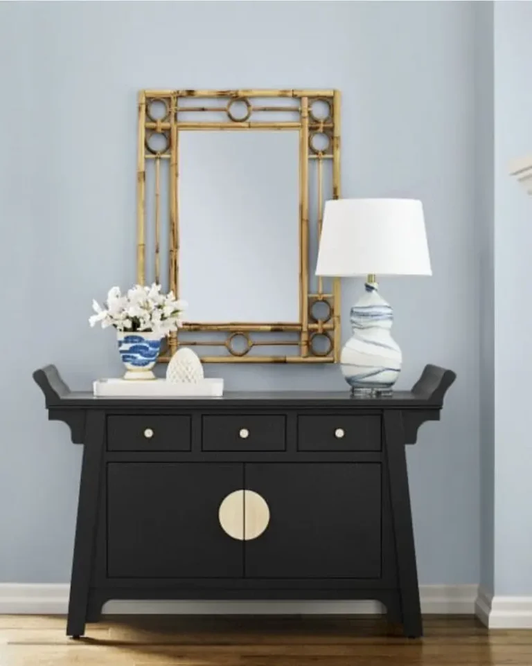 A modern-vintage black dresser with a round gold element and a blue and white lamp and flower vase on it stands against a gray-ish blue wall with a rattan mirror on it.