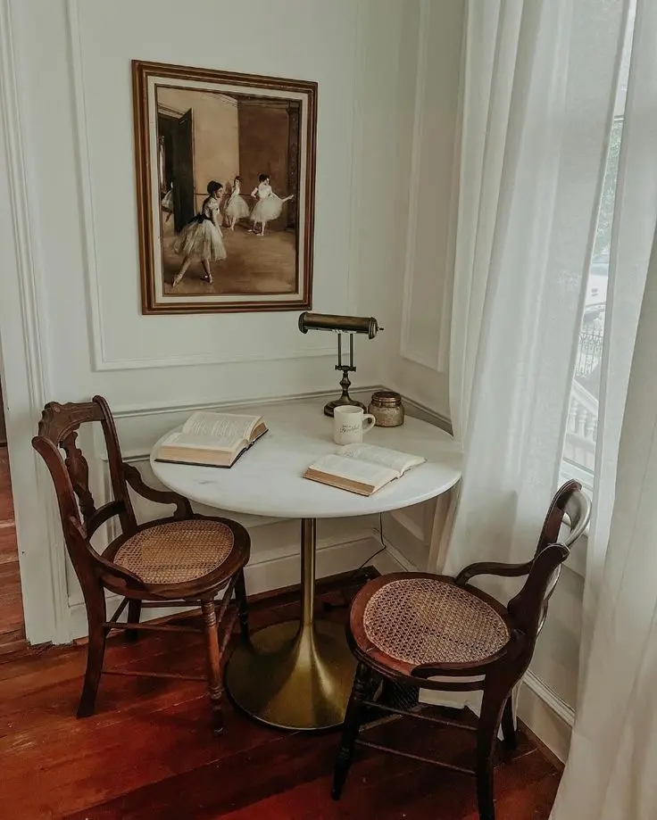 An old-fashioned nook with a wooden floor, a round marble table on brass leg, two antique cane chairs, a vintage lamp, two open books, a poster of an Edgar Degas painting, crown molding on the wall, and a long window with long white curtains.