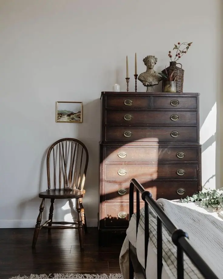 A brown wooden vintage farmhouse chair stands next to a vintage wooden dresser displaying two brass candlestick holders, a bust sculpture, and a wicker flower vase with dried flowers. There’s a white wall and a dark wood floor, and a partially visible black metal bed with a white bed throw.