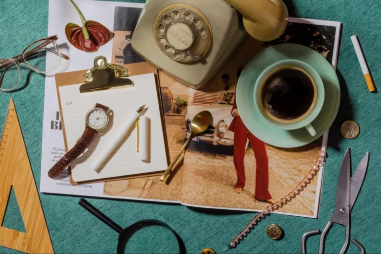 A collection of vintage office decor items set on a turquoise-colored surface: a rotary phone, wooden set square, magnifying glass, scissors, a wristwatch, glasses, a gold-tone teaspoon, fountain pen, notebook, cigarette, coins, and a cup of coffee all spread over an open magazine.