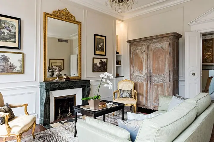 A living room with decorative molding, an ornate brass mirror, a marble fireplace mantel, two antique armchairs, a distressed antique wooden wardrobe, classical artwork on the wall, a couch, and a vintage coffee table with a potted orchid and some accessories.