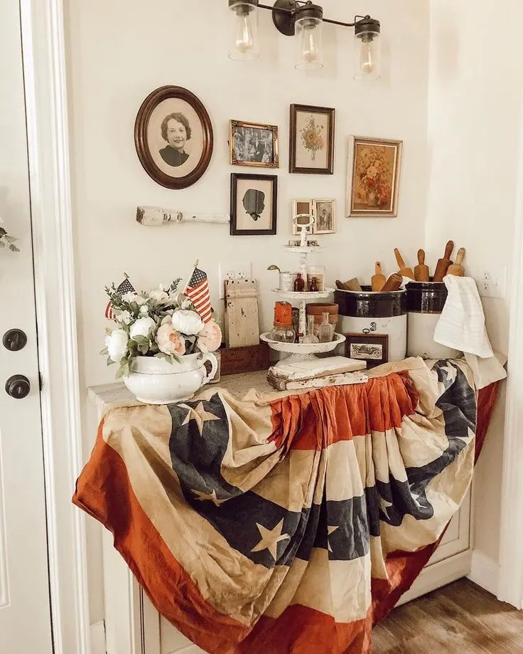 A vintage gallery wall made up of an old black and white photo in an oval frame and some botanical illustrations hangs over a vignette made up of a vase with peonies and American flags, a three-tiered distressed tray with some accessories, and two vintage crock pots with wooden rolling pins. A large vintage U.S. stars and stripes bunting hangs in the front.