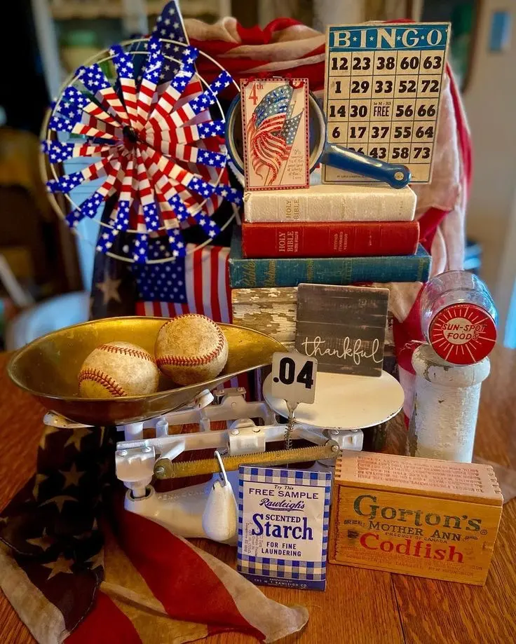 A vintage patriotic vignette featuring a stack of books, a vintage packet of starch, a retro codfish product box, an old bingo card, a vintage kitchen scale with two old baseballs, an American flag, and some patriotic flair.