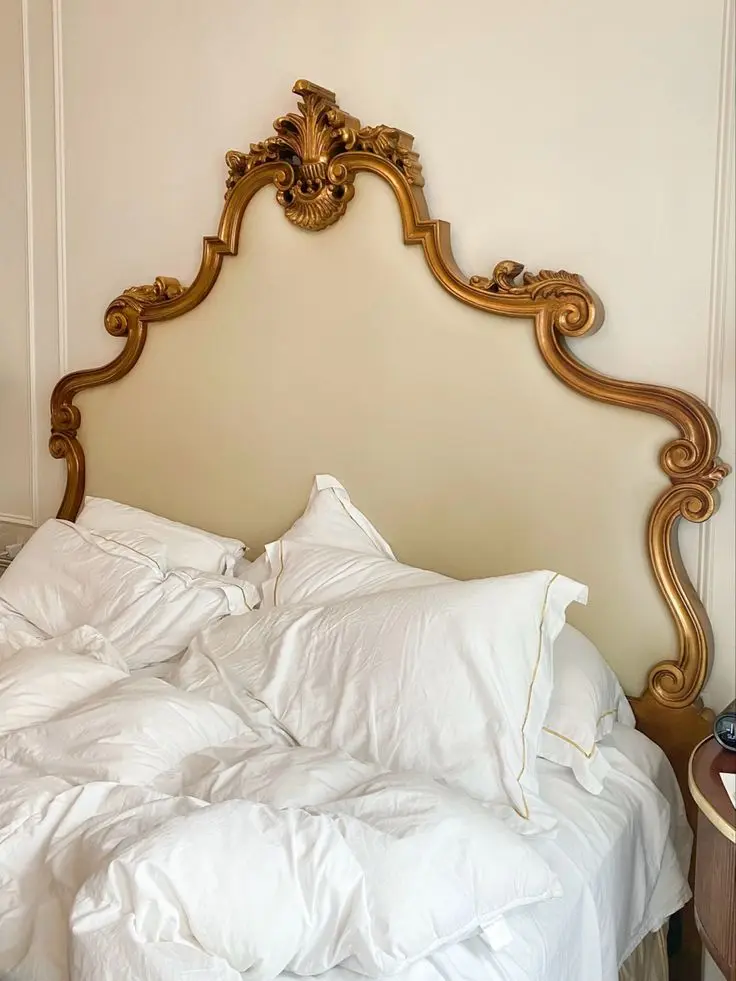 Crumpled white cotton bedding with a golden trim on a bed with a high cream colored headboard decorated with an ornate brass design.