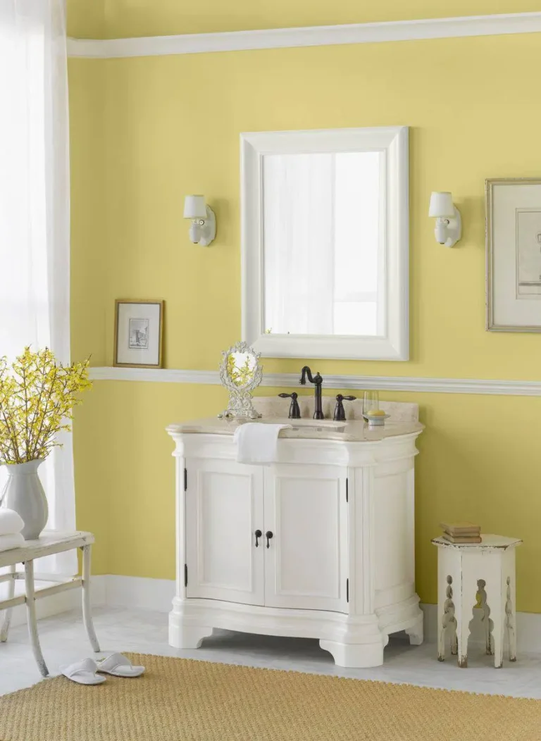 A vintage-style bathroom with yellow walls and vintage white mirror, sconces, stool, and sink dresser.