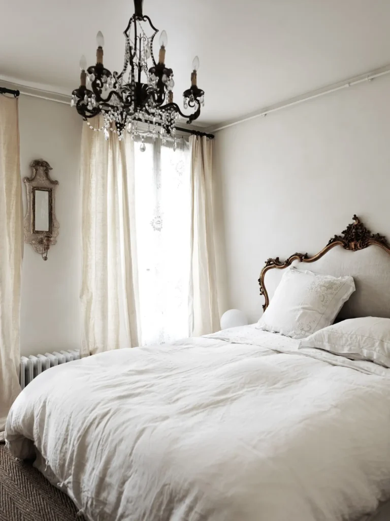 A bed with an ornate headboard and off-white linen bedding, a crystal chandelier, flowy cream curtains, white walls, an antique wall mirror.