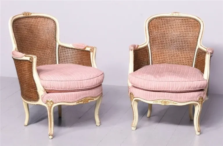 Two cream-colored Bergère chairs with cane backs, pink upholstery, and curved legs with gilded ornamental designs.