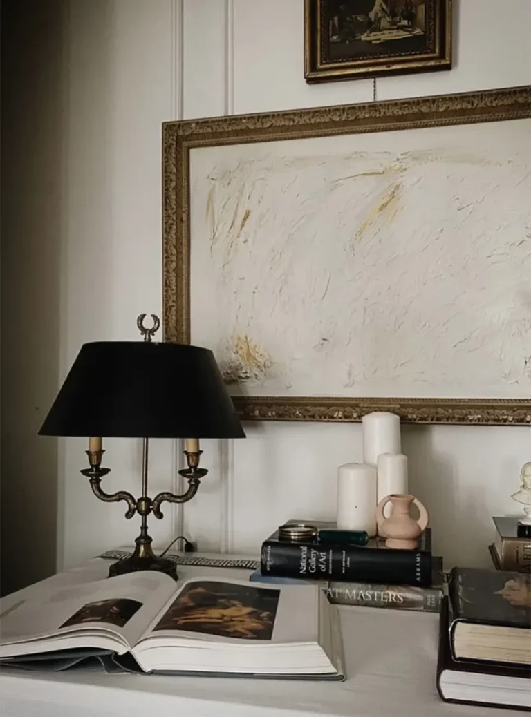 A black and brass vintage bouillotte lamp on a desk with an open book, stacks of books, three white candles, and a large ornate gilded frame on the wall.